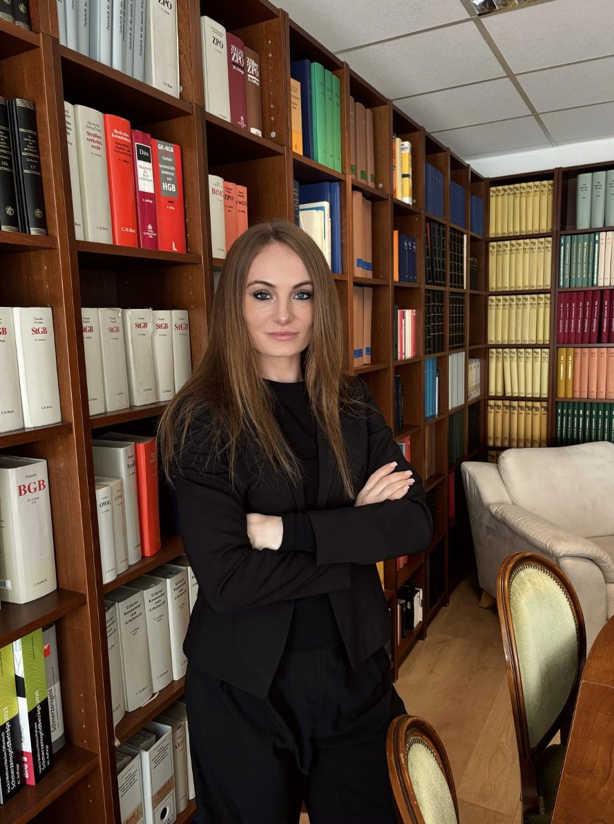 Eine Frau mit langen braunen Haaren und ernstem Blick steht vor Bücherregalen